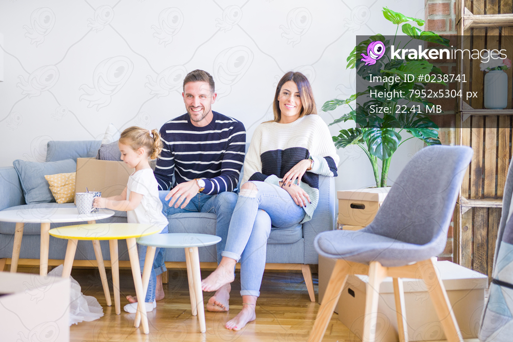Beautiful family, parents sitting on the sofa drinking coffee looking his kid playing at new home around cardboard boxes