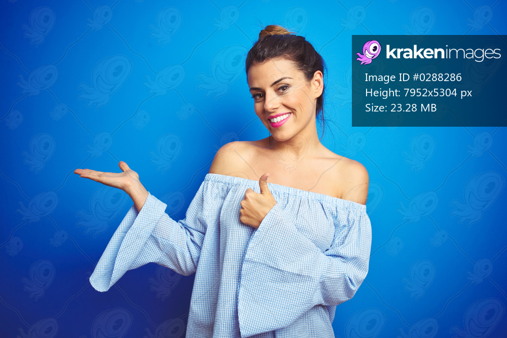 Young beautiful woman wearing bun hairstyle over blue isolated background Showing palm hand and doing ok gesture with thumbs up, smiling happy and cheerful