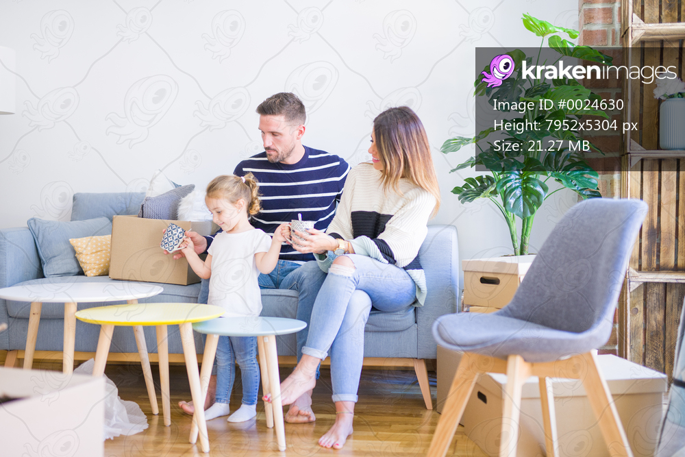 Beautiful family, parents sitting on the sofa drinking coffee looking his kid playing at new home around cardboard boxes