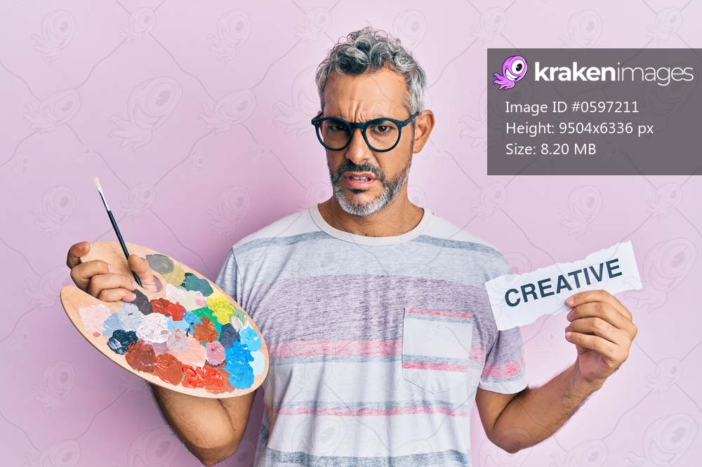 Middle age grey-haired man holding palette and creative message paper clueless and confused expression. doubt concept. 