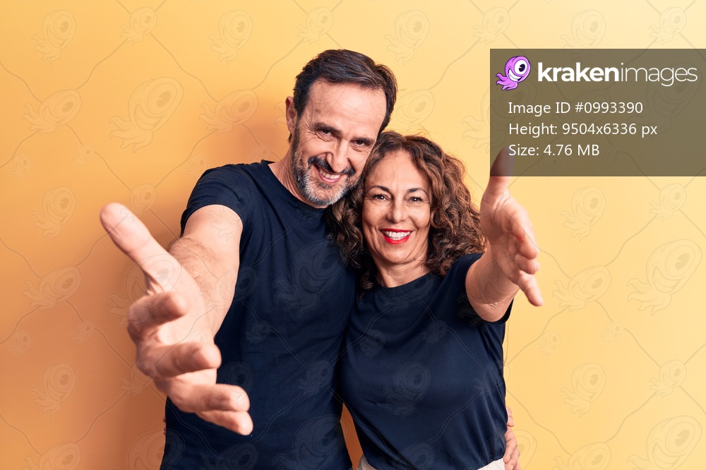 Middle age beautiful couple wearing t-shirt standing over isolated yellow background looking at the camera smiling with open arms for hug. Cheerful expression embracing happiness.