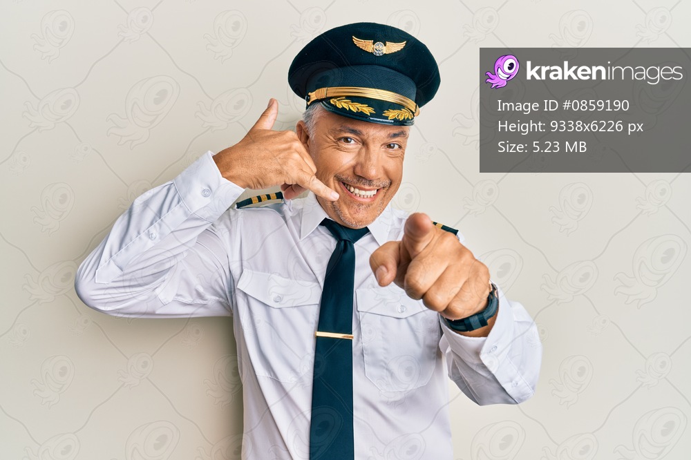 Handsome middle age mature man wearing airplane pilot uniform smiling doing talking on the telephone gesture and pointing to you. call me. 