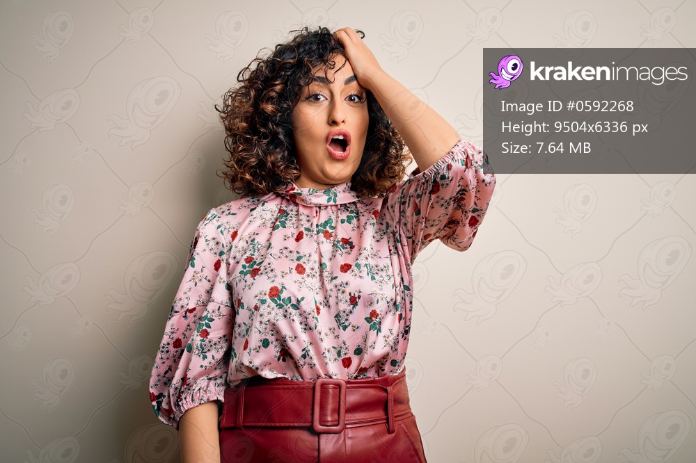Young beautiful curly arab woman wearing floral t-shirt standing over isolated white background surprised with hand on head for mistake, remember error. Forgot, bad memory concept.