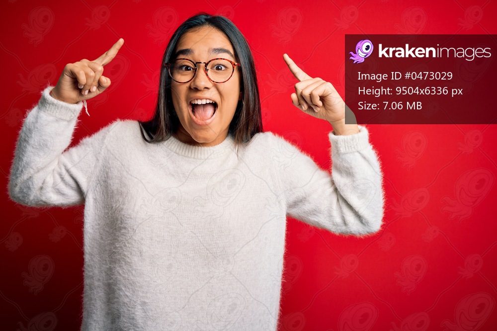 Young beautiful asian woman wearing casual sweater and glasses over red background smiling amazed and surprised and pointing up with fingers and raised arms.