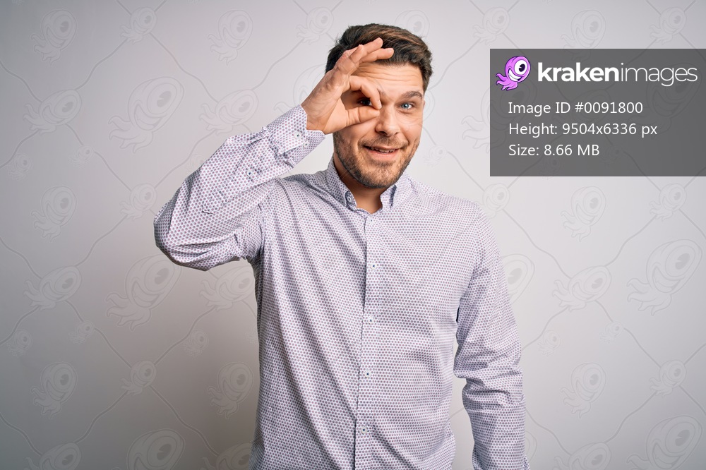 Young business man with blue eyes standing over isolated background doing ok gesture with hand smiling, eye looking through fingers with happy face.