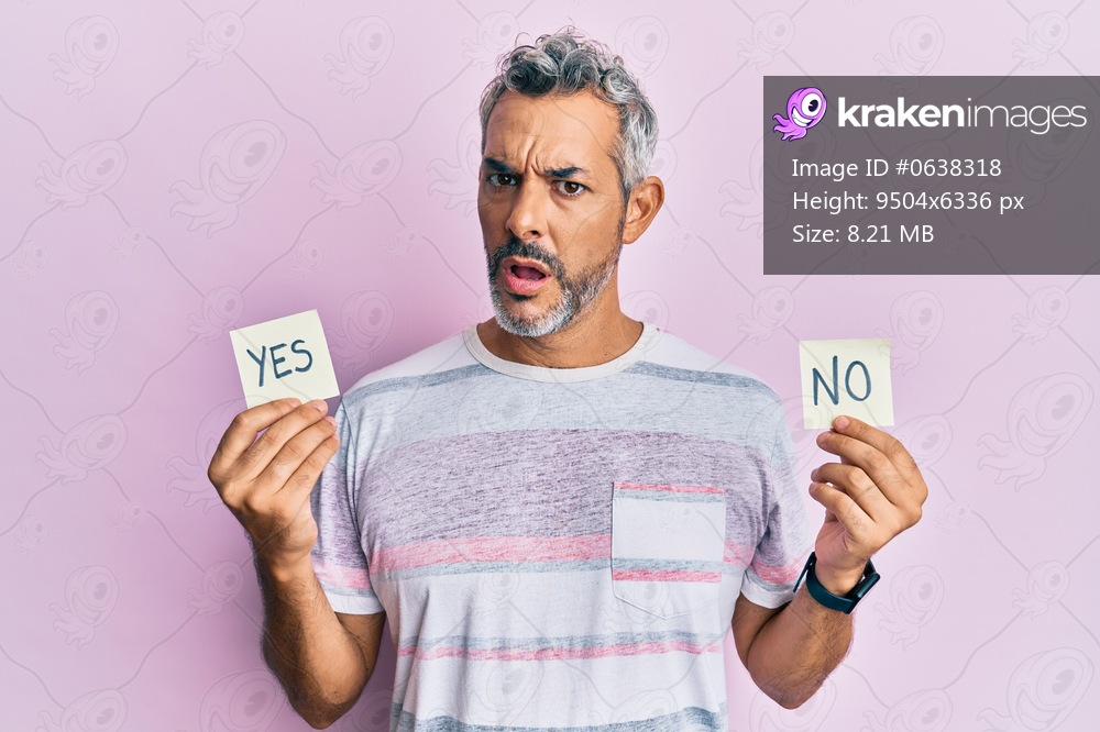 Middle age grey-haired man holding yes and no reminder in shock face, looking skeptical and sarcastic, surprised with open mouth 