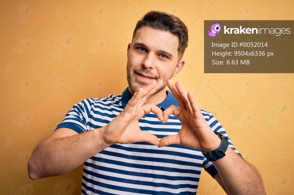 Young man with blue eyes wearing casual striped t-shirt over yellow background smiling in love showing heart symbol and shape with hands. Romantic concept.
