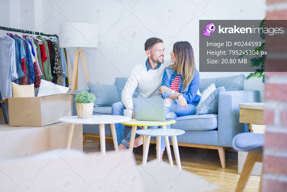 Young beautiful couple sitting on the sofa drinking cup of coffee using laptop at new home around cardboard boxes