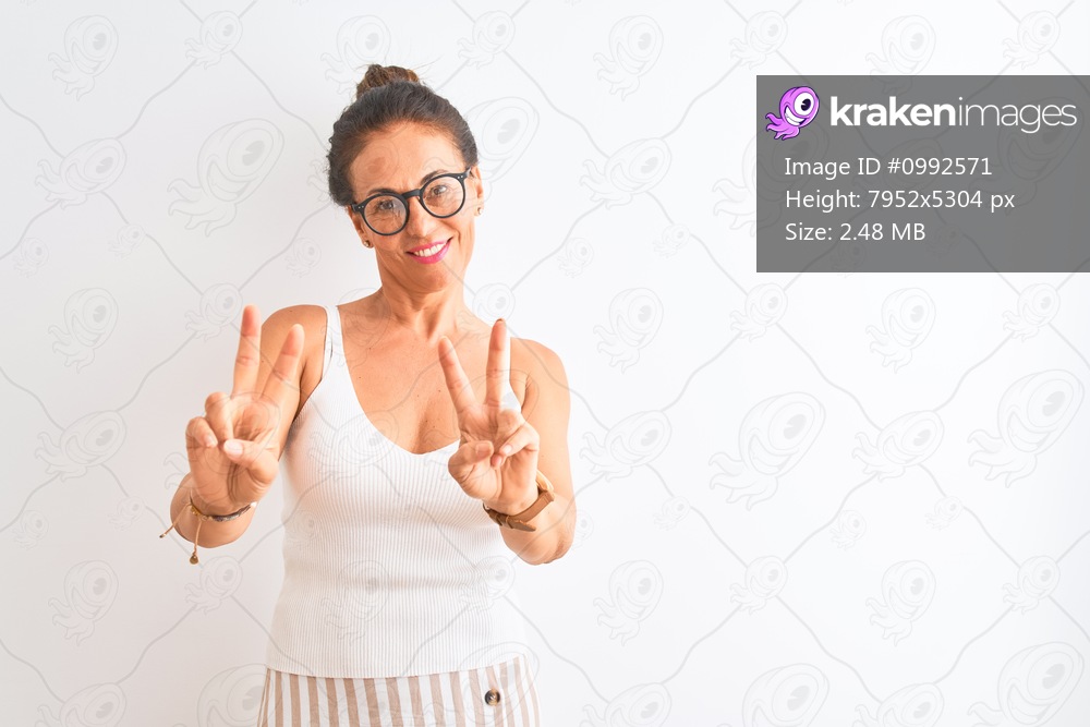 Middle age woman wearing casual t-shirt and glasses standing over isolated white background smiling looking to the camera showing fingers doing victory sign. Number two.