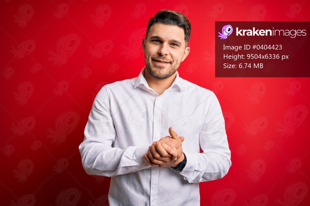 Young business man with blue eyes wearing elegant shirt standing over red isolated background with hands together and crossed fingers smiling relaxed and cheerful. Success and optimistic