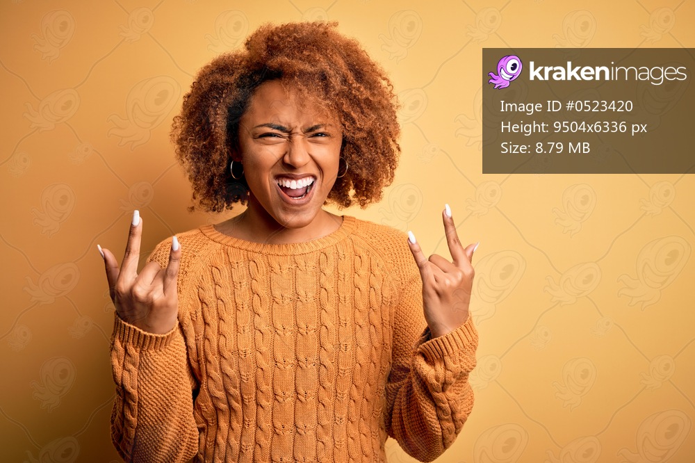 Young beautiful African American afro woman with curly hair wearing casual sweater shouting with crazy expression doing rock symbol with hands up. Music star. Heavy concept.