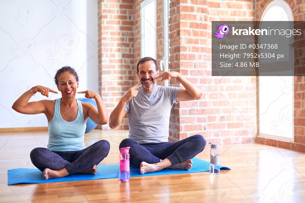 Middle age sporty couple sitting on mat doing stretching yoga exercise at gym smiling cheerful showing and pointing with fingers teeth and mouth. Dental health concept.