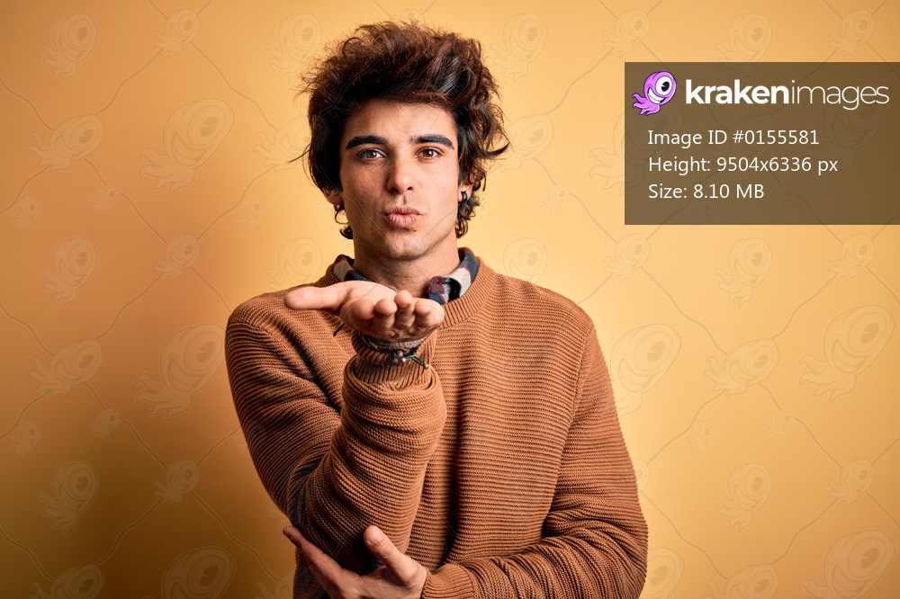 Young handsome man wearing casual shirt and sweater over isolated yellow background looking at the camera blowing a kiss with hand on air being lovely and sexy. Love expression.