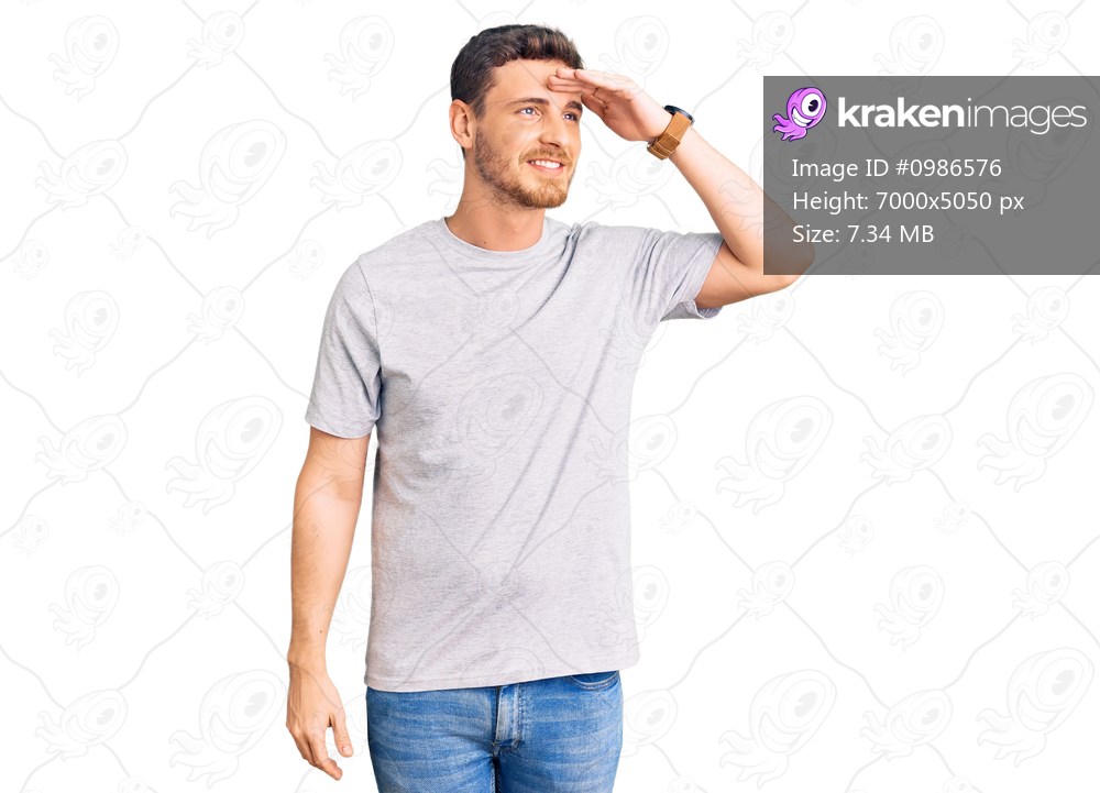 Handsome young man with bear wearing casual tshirt very happy and smiling looking far away with hand over head. searching concept. 