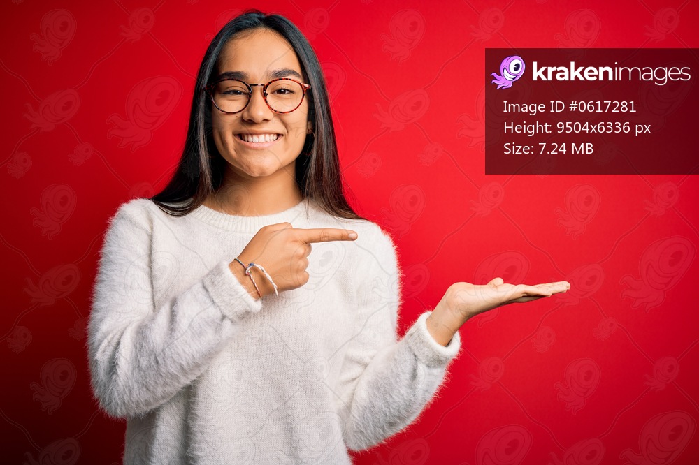 Young beautiful asian woman wearing casual sweater and glasses over red background amazed and smiling to the camera while presenting with hand and pointing with finger.
