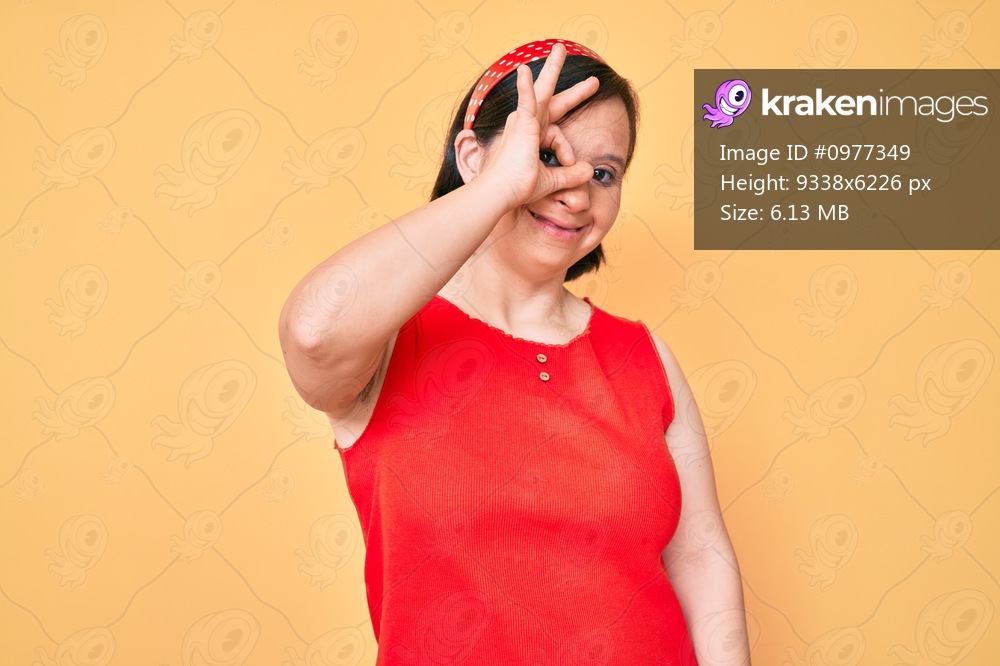 Brunette woman with down syndrome wearing casual clothes smiling happy doing ok sign with hand on eye looking through fingers 