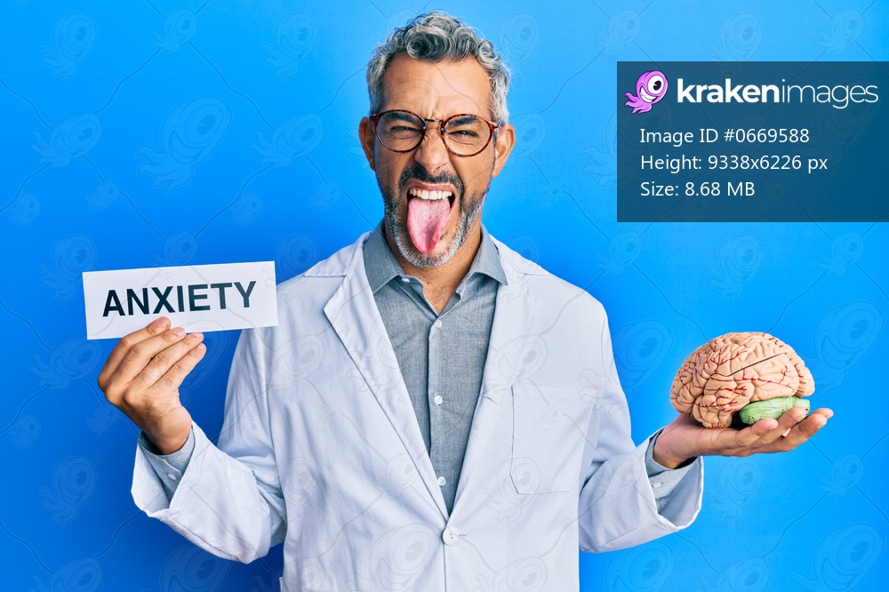 Middle age grey-haired man wearing doctor coat holding brain and anxiety message sticking tongue out happy with funny expression. 