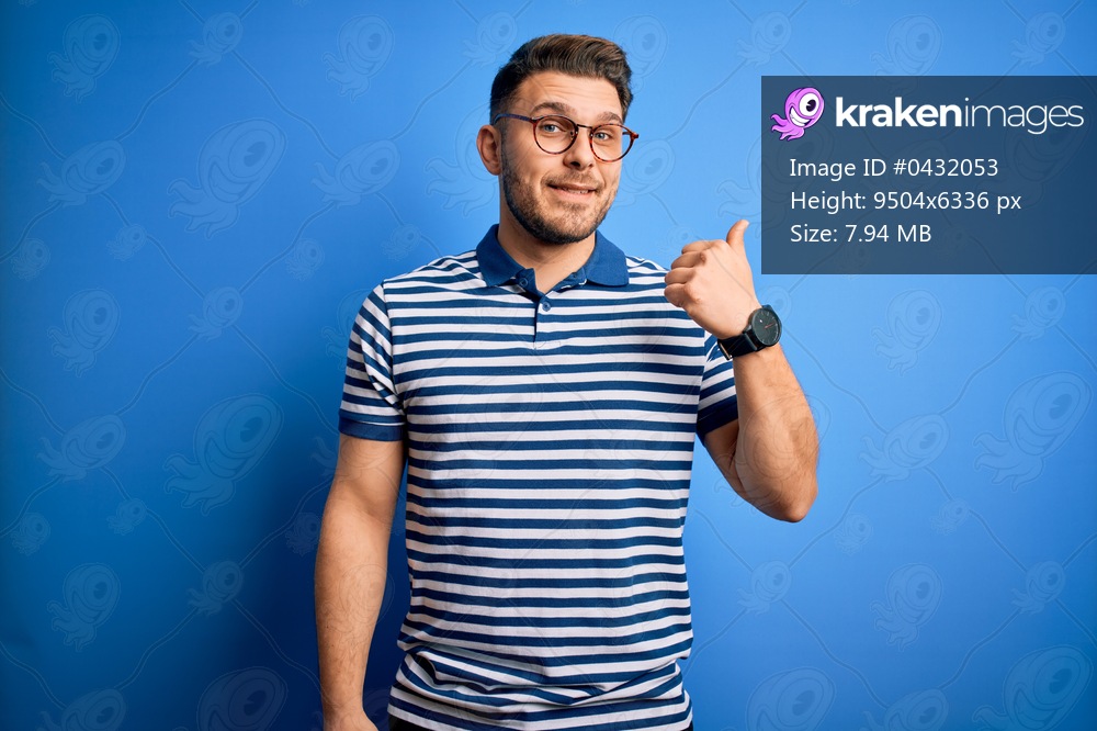 Young man with blue eyes wearing glasses and casual striped t-shirt over blue background smiling with happy face looking and pointing to the side with thumb up.