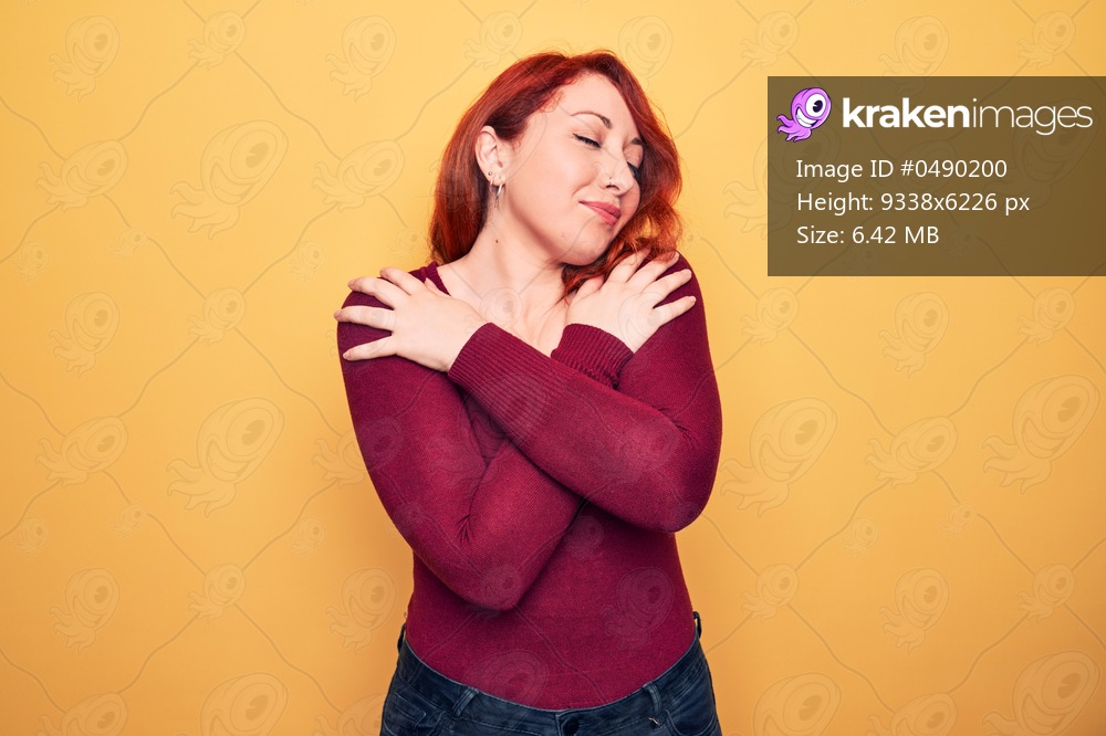 Young beautiful redhead woman wearing casual sweater standing over yellow background hugging oneself happy and positive, smiling confident. Self love and self care
