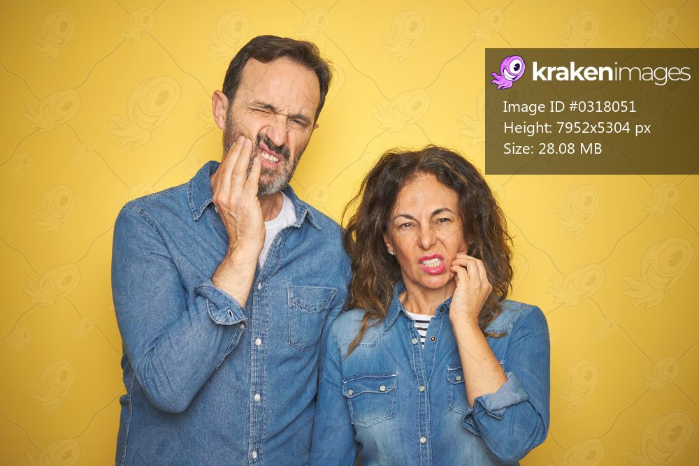 Beautiful middle age couple together standing over isolated yellow background touching mouth with hand with painful expression because of toothache or dental illness on teeth. Dentist concept.