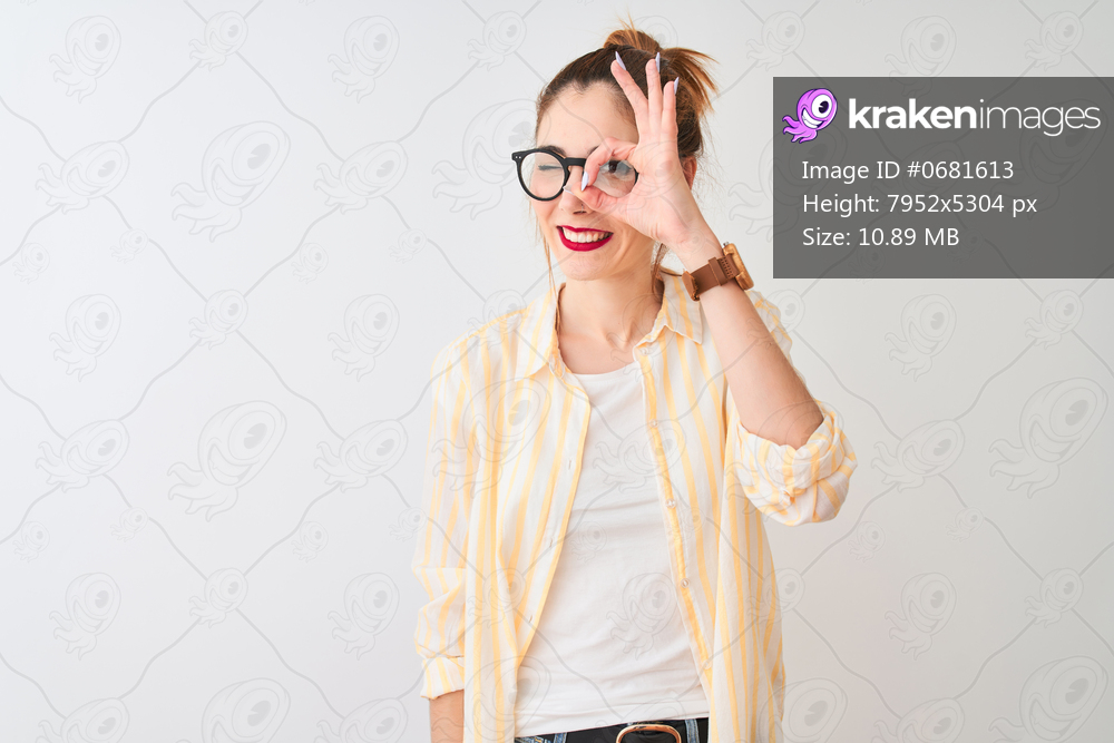 Redhead woman wearing striped shirt and glasses standing over isolated white background doing ok gesture with hand smiling, eye looking through fingers with happy face.