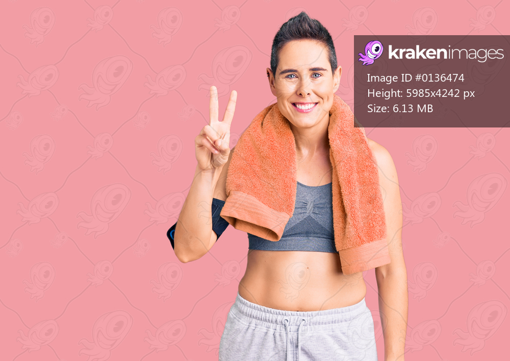 Young woman with short hair wearing sportswear and towel using smartphone showing and pointing up with fingers number two while smiling confident and happy. 
