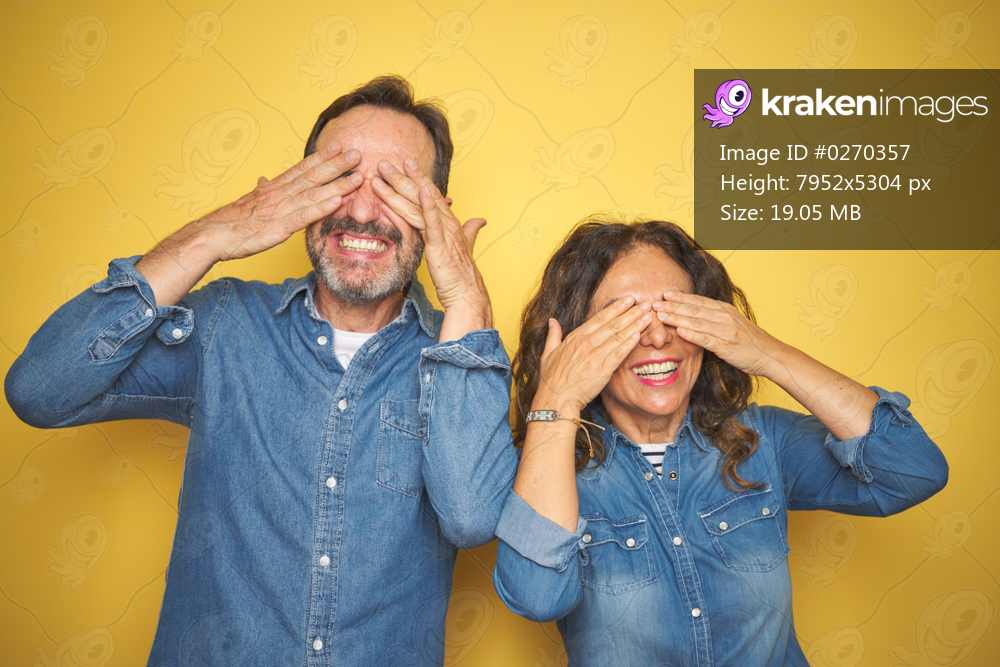 Beautiful middle age couple together standing over isolated yellow background covering eyes with hands smiling cheerful and funny. Blind concept.