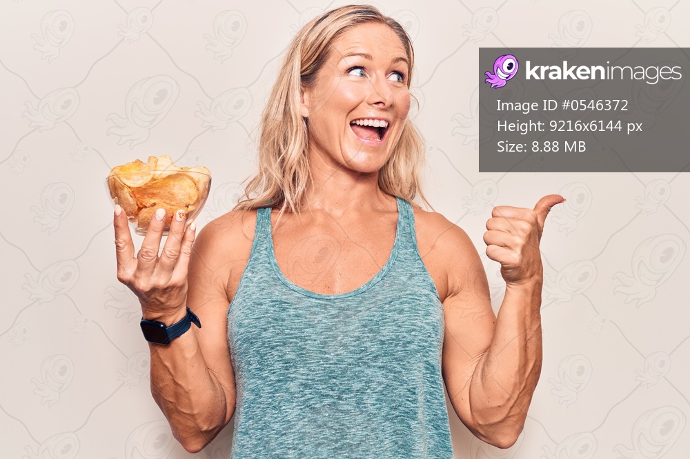 Middle age caucasian blonde woman holding potato chips pointing thumb up to the side smiling happy with open mouth 
