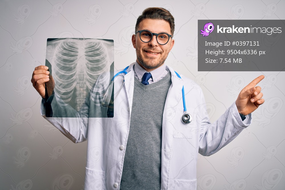 Young doctor man wearing medical coat looking at chest scan radiography over isolated background very happy pointing with hand and finger to the side