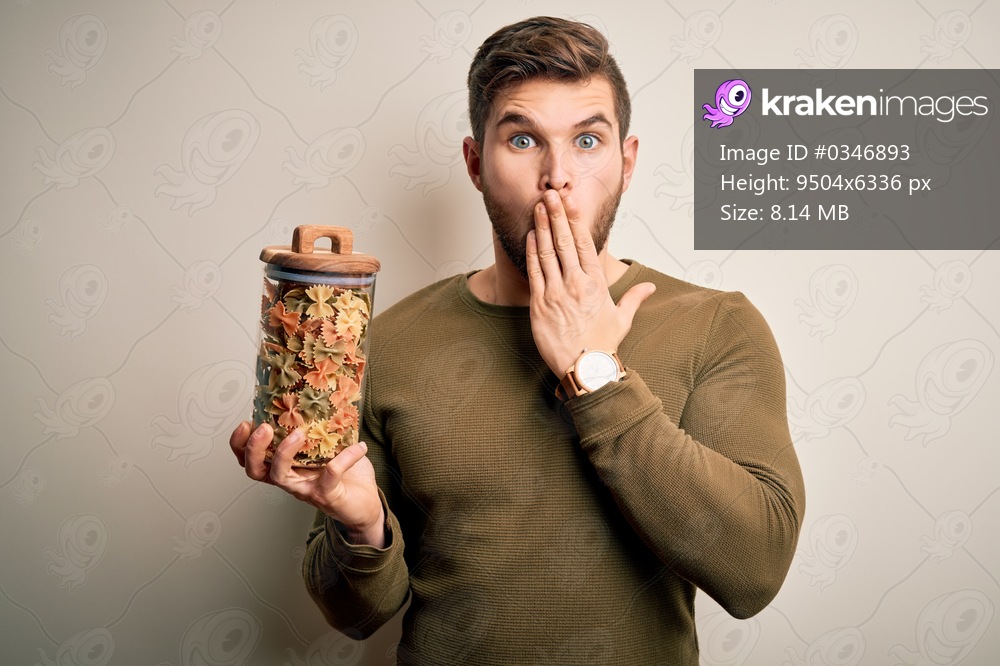 Young blond man with beard and blue eyes holding bottle of Italian dry pasta cover mouth with hand shocked with shame for mistake, expression of fear, scared in silence, secret concept