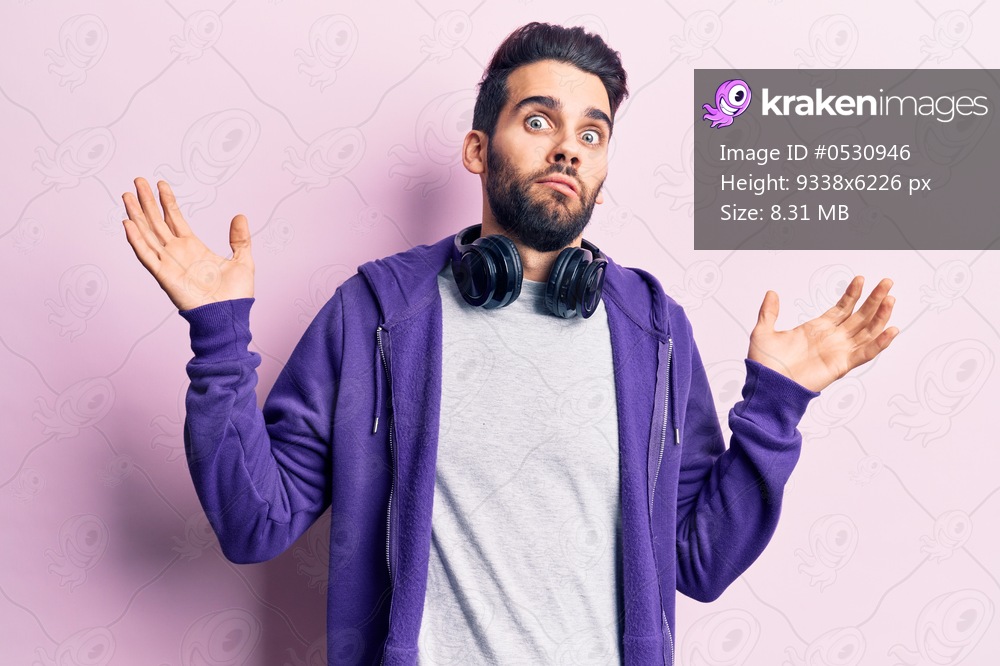 Young handsome man with beard listening to music using headphones clueless and confused with open arms, no idea and doubtful face. 