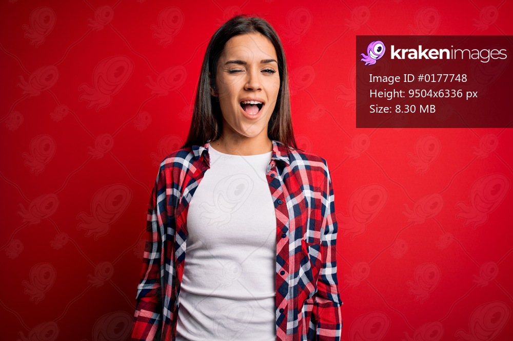 Young beautiful brunette woman wearing casual shirt standing over isolated red background winking looking at the camera with sexy expression, cheerful and happy face.