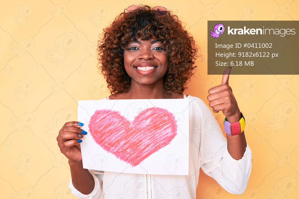 Young african american woman holding heart shape paper smiling happy and positive, thumb up doing excellent and approval sign 