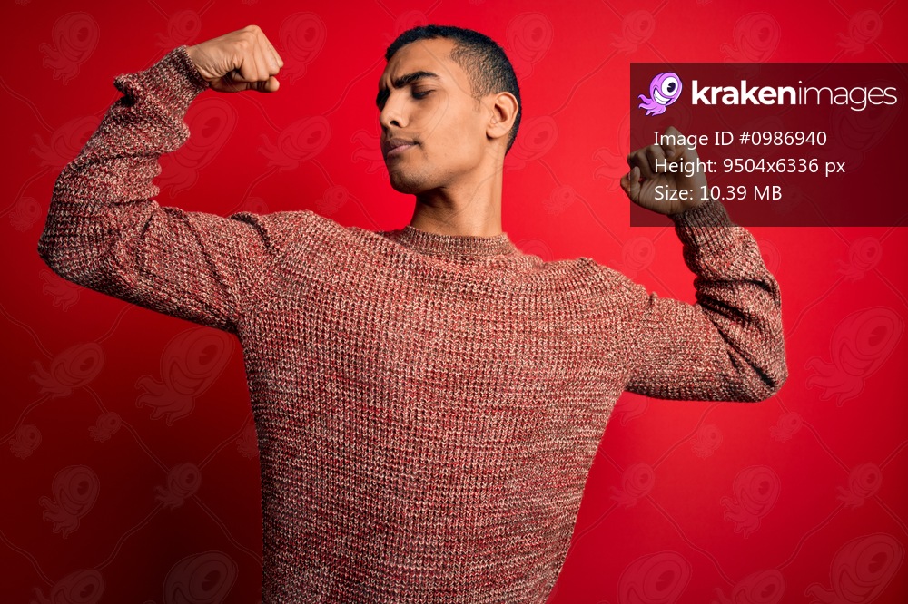 Young handsome african american man wearing casual sweater standing over red background showing arms muscles smiling proud. Fitness concept.
