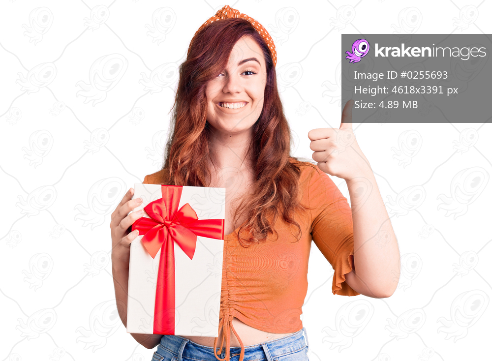 Young beautiful woman holding gift smiling happy and positive, thumb up doing excellent and approval sign 