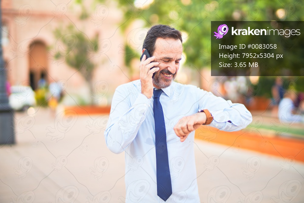 Middle age handsome businessman talking on the smartphone looking at watch smiling