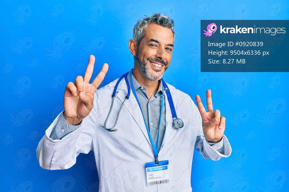 Middle age grey-haired man wearing doctor uniform and stethoscope smiling looking to the camera showing fingers doing victory sign. number two. 