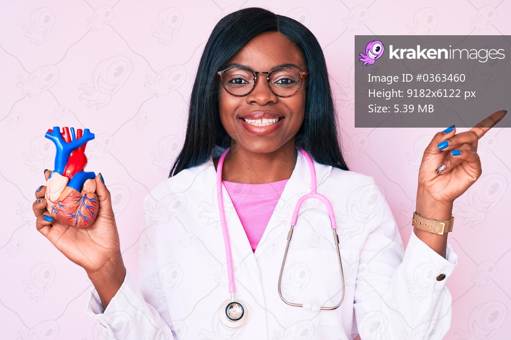 Young african american woman wearing doctor uniform holding heart smiling happy pointing with hand and finger to the side 