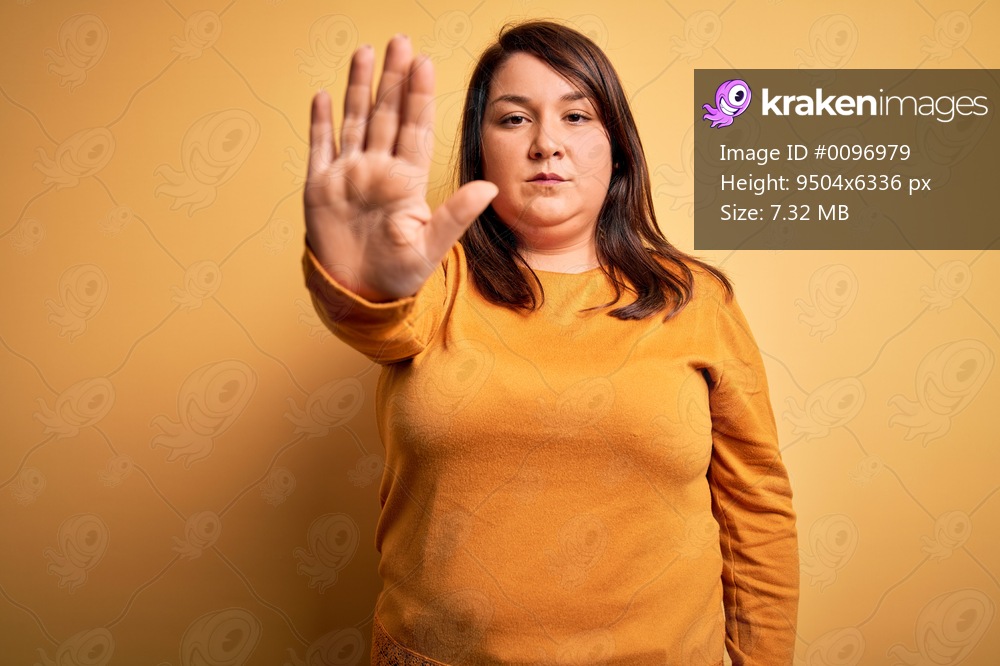 Beautiful brunette plus size woman wearing casual sweater over isolated yellow background doing stop sing with palm of the hand. Warning expression with negative and serious gesture on the face.