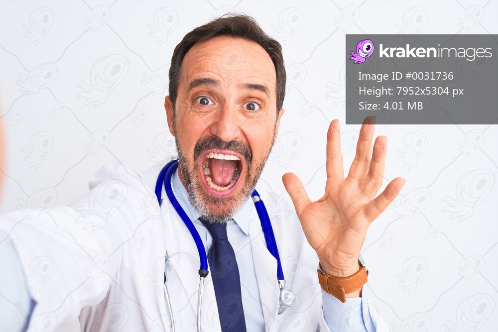 Middle age doctor man wearing stethoscope make selfie over isolated white background very happy and excited, winner expression celebrating victory screaming with big smile and raised hands