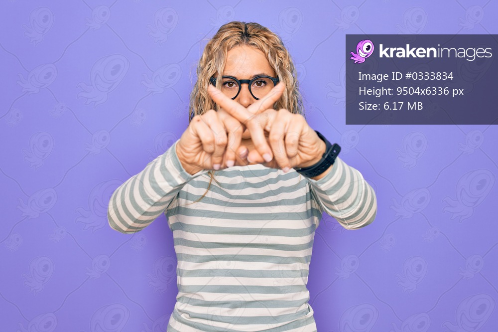 Beautiful blonde woman wearing casual striped t-shirt and glasses over purple background Rejection expression crossing fingers doing negative sign