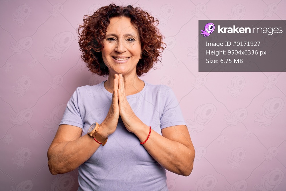 Middle age beautiful curly hair woman wearing casual t-shirt over isolated pink background praying with hands together asking for forgiveness smiling confident.