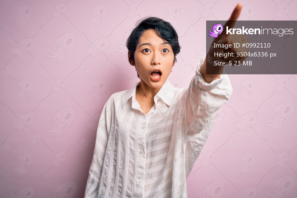 Young beautiful asian girl wearing casual shirt standing over isolated pink background Pointing with finger surprised ahead, open mouth amazed expression, something on the front