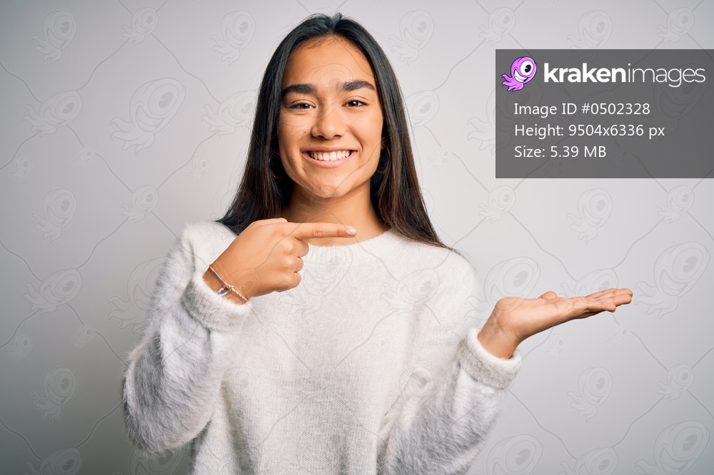 Young beautiful asian woman wearing casual sweater standing over white background amazed and smiling to the camera while presenting with hand and pointing with finger.