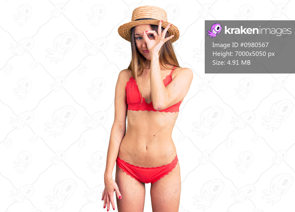 Beautiful brunette young woman wearing bikini doing ok gesture with hand smiling, eye looking through fingers with happy face. 