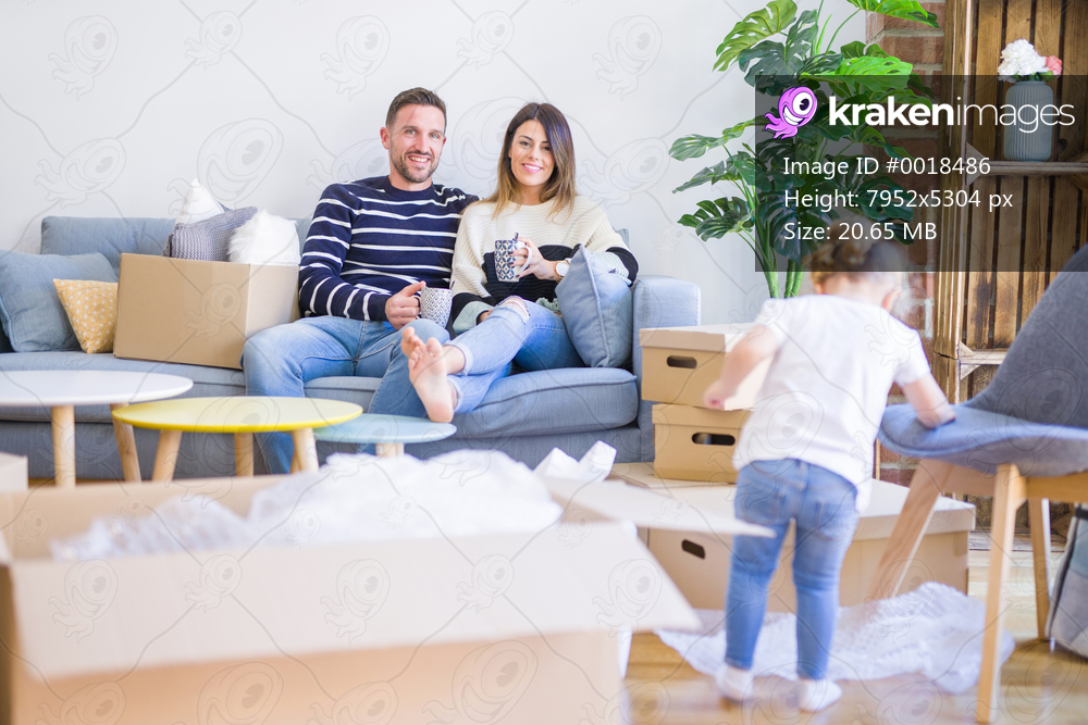 Beautiful family, parents sitting on the sofa drinking coffee looking his kid playing at new home around cardboard boxes