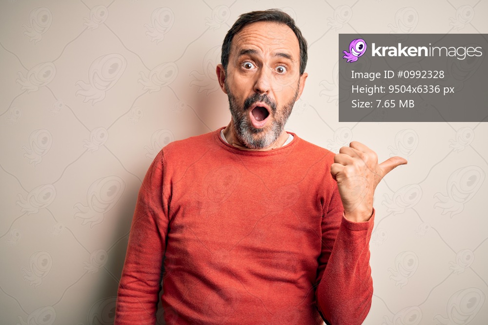 Middle age hoary man wearing casual orange sweater standing over isolated white background Surprised pointing with hand finger to the side, open mouth amazed expression.