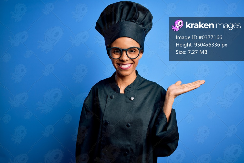 Young african american chef woman wearing cooker uniform and hat over blue background smiling cheerful presenting and pointing with palm of hand looking at the camera.