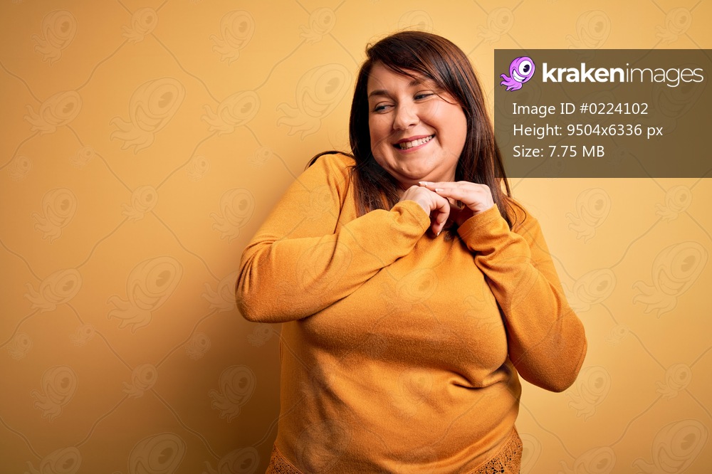 Beautiful brunette plus size woman wearing casual sweater over isolated yellow background laughing nervous and excited with hands on chin looking to the side