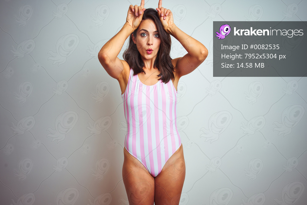 Young beautiful woman wearing striped pink swimsuit swimwear over isolated background doing funny gesture with finger over head as bull horns